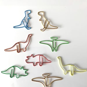 May include: A set of eight colourful dinosaur-shaped paper clips. The clips are made of metal wire and are in various colours, including blue, gold, green, red, and yellow.