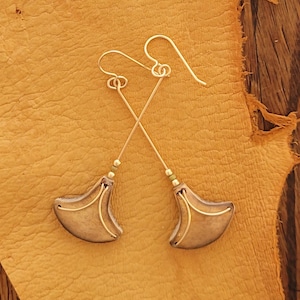 May include: A pair of handcrafted earrings with a unique design. The earrings feature a gold-colored hook and a long, thin gold wire that crosses another wire. The bottom of the earrings have a bronze-colored, crescent-shaped design with gold wire accents.