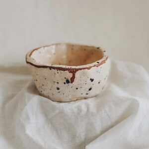 May include: A small, hand-formed ceramic bowl with an irregular, organic shape. The bowl features a cream-colored base with scattered black speckles and a distinct, uneven reddish-brown rim that drips slightly down the exterior side of the vessel.