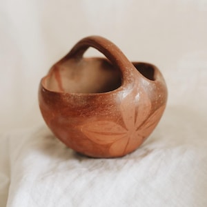 May include: A small, round, terracotta-colored ceramic basket with a handle. The basket features a floral design carved into the side. The handle arches over the top, and the interior is unglazed. The basket sits on a white, textured surface.