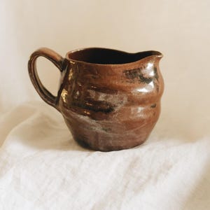 May include: A small, hand-thrown ceramic creamer pitcher with a glossy, dark brown glaze and subtle black accents. The vessel features an organic, slightly uneven shape, a curved handle on the side, and a small, integrated pouring spout at the rim.