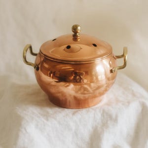 May include: A polished copper pot with a rounded body, two brass side handles, and a matching lid featuring a small brass knob. The lid has several small circular vents along the top edge, and the entire piece has a reflective metallic finish.