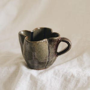 May include: A ceramic mug with a flower-shaped rim and a curved handle. The mug features a mottled, earthy glaze in shades of olive green, brown, and dark grey, with a textured surface and visible vertical indentations along the exterior body.