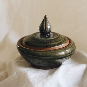 May include: A round, lidded ceramic jar featuring a glossy, dark olive-green glaze over a terracotta-coloured clay base. The lid is topped with a small, pointed, teardrop-shaped handle, and the overall design showcases a smooth, hand-thrown finish.