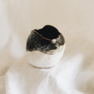 May include: A small, rounded ceramic vessel with an irregular, organic rim. The upper portion features a glossy, dark charcoal-black glaze, while the lower section is finished in a matte, off-white glaze, creating a distinct two-tone appearance.
