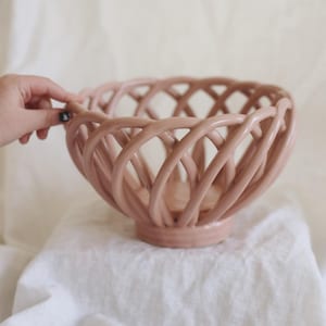 May include: A blush pink ceramic bowl with a woven design. The bowl has a round base and a lattice-like pattern around the sides. The bowl is sitting on a white surface.