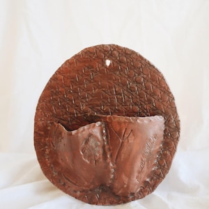 May include: A brown, handcrafted wall pocket with a woven texture on the back and a pocket with leaf details. The wall pocket is made of a dark brown material and has a hole for hanging. The overall shape is oval.