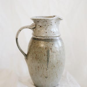 May include: A handmade ceramic pitcher with a speckled, off-white rim and handle. The body of the pitcher is a muted green-gray with subtle brown speckles. The pitcher has a classic, rounded shape and a ruffled rim.