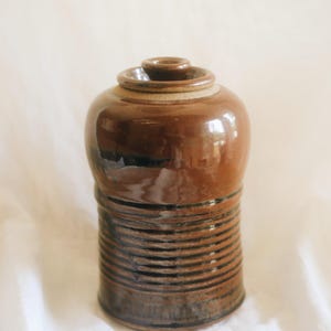 May include: A brown ceramic jar with a glossy finish and a matching lid. The upper portion of the jar is smooth and rounded, while the lower section features a series of horizontal ridges and dark brown to black linear detailing throughout.
