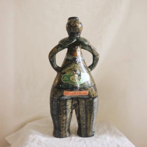 May include: A ceramic bottle shaped like a stylized figure with hands on hips, featuring a mottled green, brown, and black glaze. The bottle has a small neck and two labels, one green with the text ART and one orange with text including SAN FRANCISCO.