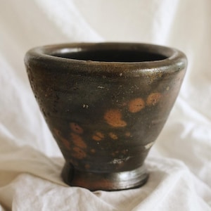 May include: A dark brown, handcrafted mortar with a wide mouth and a flared shape. The mortar has a textured surface with lighter brown spots and sits on a small, circular base. The background is a white, wrinkled fabric.