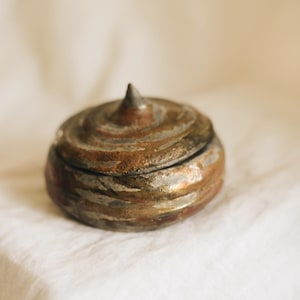 May include: A small, round, lidded ceramic container with a textured, metallic-toned finish in shades of bronze, copper, and dark gray. The lid features a prominent, pointed conical knob at the center, and the surface displays concentric ridges.