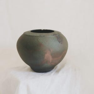 May include: A round, dark green ceramic vase with a wide opening. The vase has a textured surface with hints of reddish-brown. The vase is sitting on a white surface. The vase is approximately 6 inches in diameter and 5 inches tall.