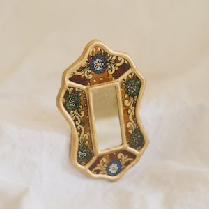 Puede incluir: Un peque&ntilde;o espejo decorativo con un marco dorado. El marco tiene una forma irregular y est&aacute; adornado con un estampado floral en tonos marr&oacute;n, azul y verde. El espejo es rectangular.