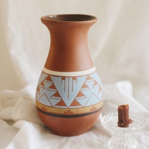 May include: A terracotta vase with a flared rim and a matte finish, featuring a painted geometric pattern in light blue, white, and gold bands around the center. Beside it sits a small, clear glass candle holder with a short, dark brown candle.