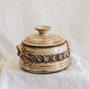 May include: A beige ceramic lidded jar with a decorative band of floral-like designs. The jar has a round shape with small handles and a small knob on the lid. The ceramic has a speckled, aged appearance.