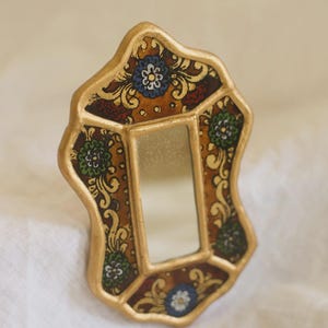 Puede incluir: Un peque&ntilde;o espejo ornamentado con un marco dorado. El marco presenta un patr&oacute;n decorativo con motivos florales en tonos marr&oacute;n, rojo, verde y azul. El espejo es rectangular y est&aacute; dentro del marco decorativo.