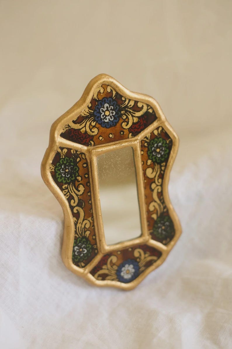 Puede incluir: Un peque&ntilde;o espejo ornamentado con un marco dorado. El marco presenta un patr&oacute;n decorativo con motivos florales en tonos marr&oacute;n, rojo, verde y azul. El espejo es rectangular y est&aacute; dentro del marco decorativo.
