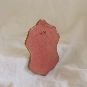 Puede incluir: Un objeto peque&ntilde;o, de forma irregular, con una superficie rosa y bordes dorados. La parte posterior del objeto tiene un peque&ntilde;o gancho de metal. El objeto descansa sobre una superficie blanca y texturizada.
