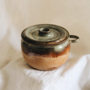 May include: A round ceramic pot with a lid and a small side handle. The pot features a two-tone glaze, with a smooth, light tan base transitioning into a dark brown and muted green drip pattern on the upper portion and the lid.