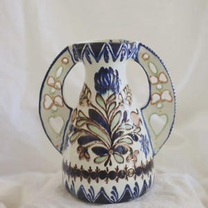 May include: A ceramic vase with a cream-colored base, featuring two large, open-work handles with heart-shaped cutouts. The body is decorated with hand-painted floral motifs in shades of dark blue, sage green, and soft brown, with a zigzag pattern.