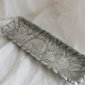 May include: A rectangular, silver-colored decorative tray with a detailed relief of rabbits nestled among leaves. The tray is made of metal and has a slightly textured surface. The design features several rabbits in various poses.