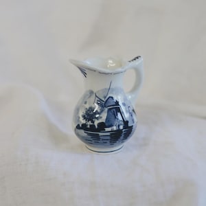 May include: A small white ceramic creamer pitcher featuring a blue hand-painted design of a traditional windmill landscape. The pitcher has a curved handle, a pouring spout, and a rounded body decorated with blue ink illustrations of a mill.