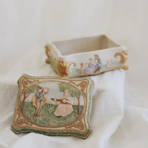 Peut inclure: Boîte rectangulaire en porcelaine avec couvercle. Le couvercle présente une scène en relief de personnages dans un jardin, encadrée d'une bordure verte et dorée. La boîte et le couvercle sont crème avec des détails dorés.