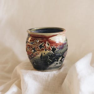 May include: A ceramic vessel featuring a cut-out design of stars and circular shapes. The exterior glaze displays a blend of deep blue, dark purple, and reddish-brown tones, while the interior rim shows a smooth, glossy finish in shades of blue.