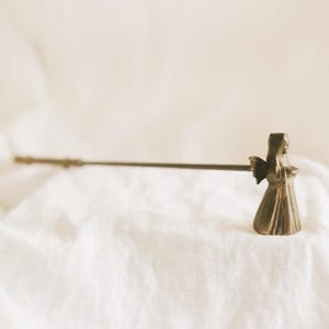 May include: A metallic bronze-toned candle snuffer featuring a sculpted angel figure at the end of a long, slender handle. The angel is depicted with folded wings and a draped gown, attached to a straight metal rod used for extinguishing candles.