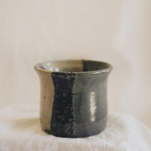 May include: A ceramic cup with a flared rim and a cylindrical body. The exterior features a two-tone glaze, with a smooth light beige section on one side and a dark, textured charcoal grey section on the other, separated by a vertical black stripe.