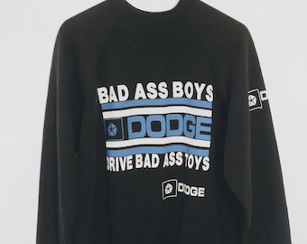 Puede incluir: Una sudadera negra de manga larga con texto gráfico en blanco y azul claro que dice BAD ASS BOYS DODGE DRIVE BAD ASS TOYS. El diseño incluye el logotipo pentastar de Dodge impreso en el frente, con un pequeño logotipo en la manga izquierda.
