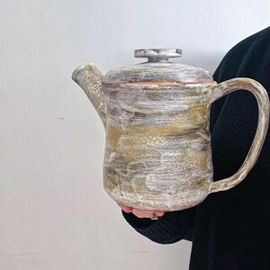 May include: A ceramic teapot with a speckled glaze in shades of brown, white, and gray. The teapot has a rounded body and a lid with a knob.