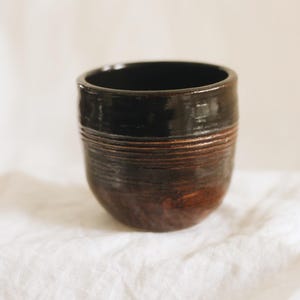 May include: A small, rounded ceramic cup featuring a glossy black glaze on the upper portion and a textured, dark brown finish on the lower body. The exterior surface displays horizontal grooved lines encircling the middle section of the vessel.
