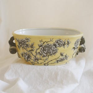 May include: A yellow ceramic bowl featuring a black floral pattern of blooming flowers and branches. The bowl has a smooth white interior and two dark, sculpted handles on opposite sides, providing a contrast to the yellow exterior surface.