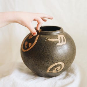 May include: A dark brown ceramic vase with a round shape and a wide opening. The vase is decorated with tan-colored abstract designs. A hand is reaching towards the top of the vase. The vase is sitting on a white cloth.