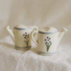 May include: Two small, cream-colored ceramic watering cans featuring a hand-painted blue floral design on the front. Each piece has a light blue horizontal band around the top and bottom, a curved handle, and a small spout extending to the side.