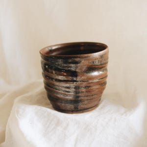 May include: A cylindrical ceramic cup featuring a dark brown and black mottled glaze with a series of horizontal ridges along the exterior surface. The rim is smooth and circular, leading down to a slightly tapered base with a visible clay texture.