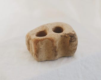 Puede incluir: Un pequeño objeto de cerámica beige de forma irregular con una superficie mate y texturizada. La pieza presenta dos aberturas circulares en la parte superior, un perfil exterior ondulado e irregular y motas de color marrón oscuro.