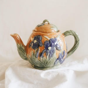 May include: A ceramic teapot with a warm orange glaze, adorned with blue iris flowers and green leaves. The handle and lid are also green. The teapot is a decorative item for serving tea.