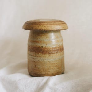 May include: A cylindrical ceramic jar with a wide, flat lid, featuring a speckled tan and brown glaze with horizontal drip patterns. The vessel has a smooth, rounded top and a sturdy base, showcasing natural earthy tones and a textured finish.