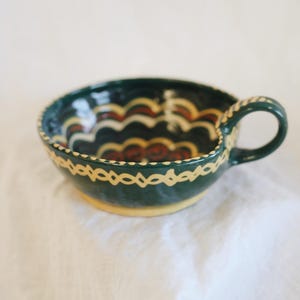 May include: A handcrafted ceramic bowl with a handle, featuring a dark green exterior and a yellow base. The interior showcases a wave-like pattern in red, yellow, and white. The rim is adorned with a decorative yellow design.