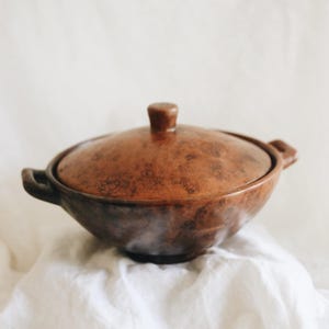 May include: A round, brown, mottled ceramic cooking pot with a matching lid and two side handles. The pot features a dark, uneven glaze with a textured, earthy appearance and a small, knob-shaped handle centered on the top of the lid.