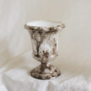 May include: A pedestal vase featuring a marbled pattern of dark brown and white swirls. The vessel has a wide, flared rim, a rounded body that tapers toward a narrow stem, and a circular base. The interior of the vase is a solid white color.