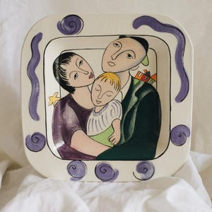 May include: Square ceramic decorative plate with a family portrait illustration. The artwork features a mother, father, and child in a stylized art deco design. The plate has a white background with purple accents and a purple border.