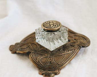 Puede incluir: Juego de tintero antiguo. El tintero de cristal transparente tiene un diseño en espiral y una tapa decorativa. Se encuentra sobre una bandeja de color bronce con un diseño ornamentado de estilo Art Nouveau. El conjunto se exhibe sobre una superficie blanca.