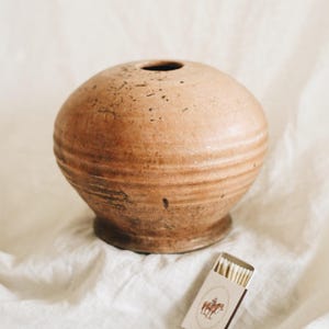 May include: A round, brown ceramic vase with a small opening at the top. The vase has horizontal ridges and sits on a small base. A matchbox with a cowboy illustration is next to the vase.