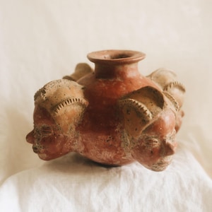 May include: A terracotta-colored ceramic vessel featuring a central circular opening and four sculpted human heads protruding from the sides. Each head displays detailed facial features and a textured, light-colored headdress with linear carvings.