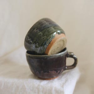 May include: Two stacked ceramic cups with a glossy, dark glaze in shades of deep purple, green, and black. The bottom cup features a small handle, while the top cup is inverted, revealing a raw, unglazed tan clay base at the bottom of the vessel.