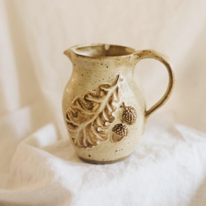 May include: A cream-colored ceramic pitcher with a speckled finish, featuring a raised relief design of a large oak leaf and two small acorns on the front. The pitcher has a curved handle and a small pouring spout at the top rim.